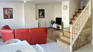 Un salon avec un canapé rouge, une table blanche et un téléviseur posé sur un meuble. On y trouve un escalier avec une rampe blanche qui mène à l’étage supérieur.