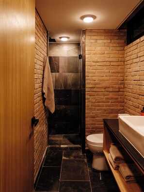 A compact bathroom with a wooden door, a glass shower enclosure, a white toilet, and a wooden vanity with a sink. - Mana by Pardela (Guadalajara)