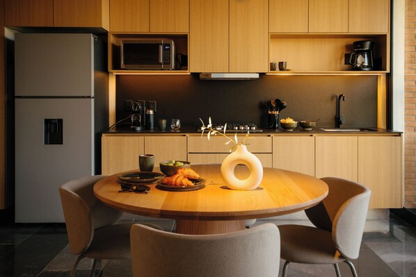 A modern kitchen with wooden cabinets, a built-in microwave, a coffee machine, and a round dining table with chairs. - Mana by Pardela (Guadalajara)