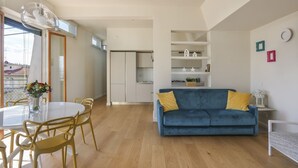 Ein modernes Wohnzimmer mit einem blauen Sofa, gelben Sesseln, einem weißen Esstisch und einer Küche im Hintergrund.
