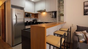 Cocina compacta con una isla central, refrigerador, microondas y cafetera. Hay dos taburetes de bar y un sofá con cojines.