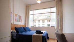 Un dormitorio con cama, una silla, ventilador de techo, ventana con cortinas y una obra de arte en la pared.