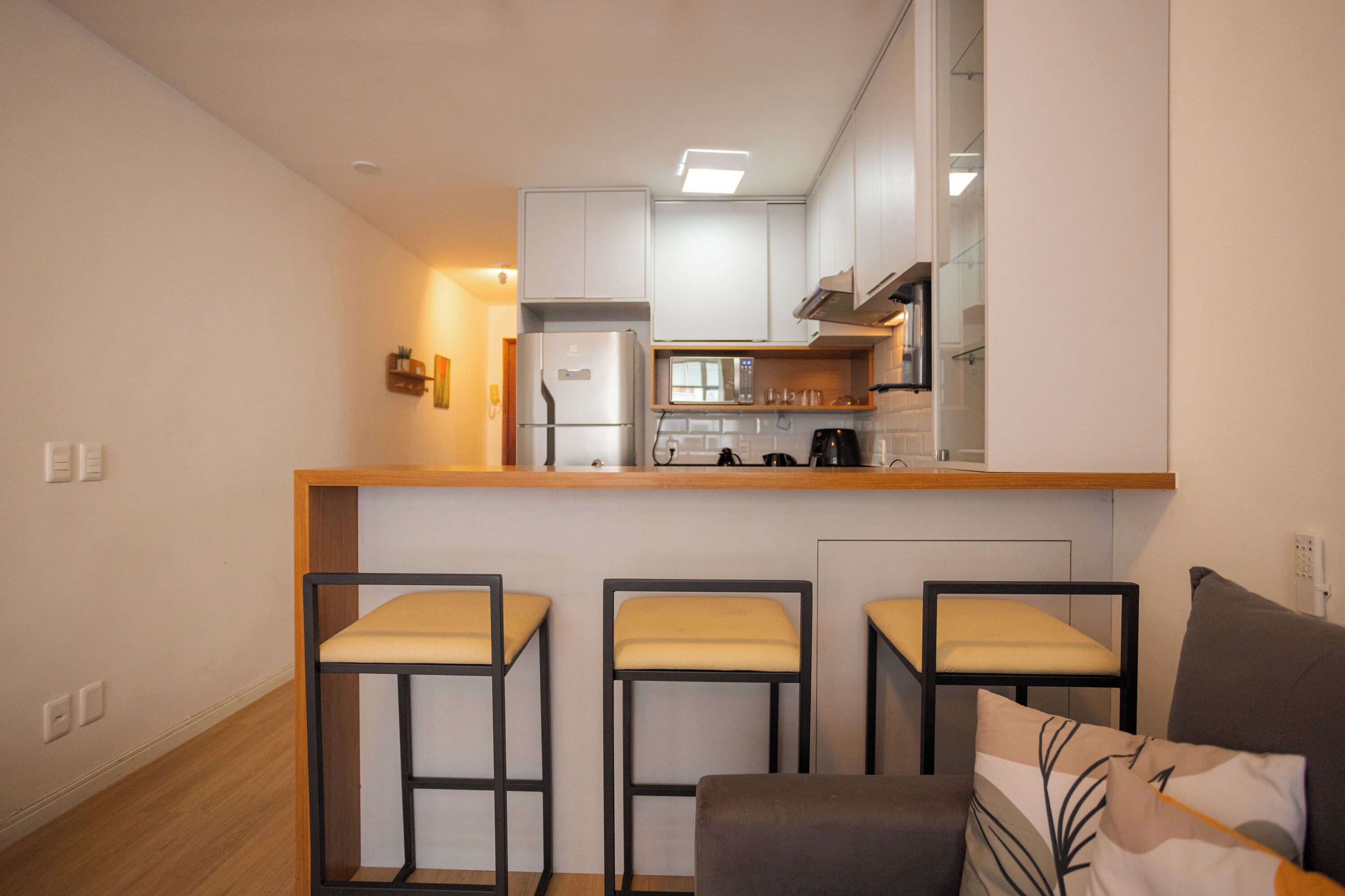 A compact kitchen with a refrigerator, microwave, and coffee maker. There are three bar stools and a sofa with a cushion.