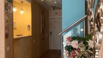 A hotel reception area with a wooden floor, a reception desk, a staircase with a glass railing, and a decorative vase with flowers.