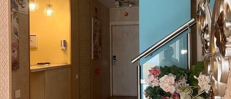 A hotel reception area with a wooden floor, a reception desk, a staircase with a glass railing, and a decorative vase with flowers.