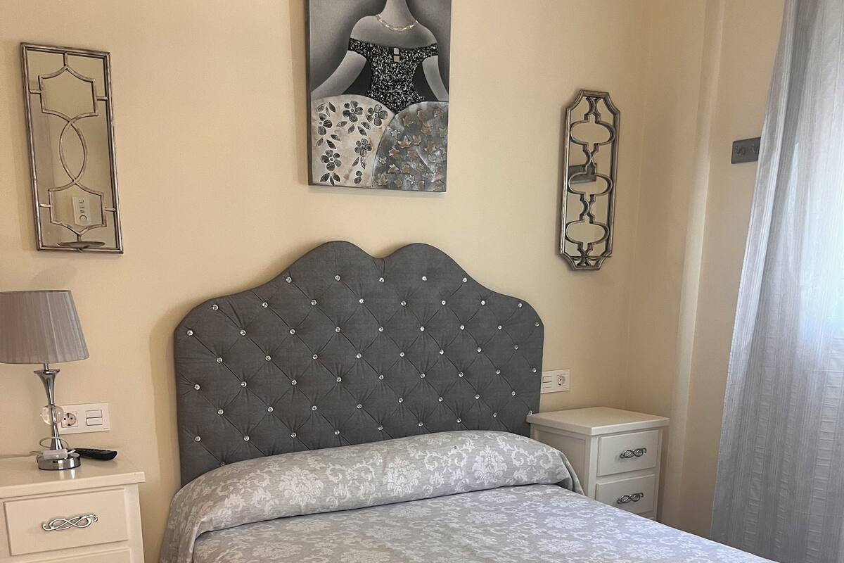 A bedroom with a tufted headboard, a patterned bedspread, a bedside table with a lamp, and a decorative mirror.