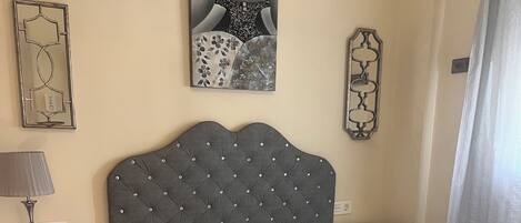Un dormitorio con cabecera capitonada, colcha estampada, mesita de noche con lámpara y espejo decorativo.