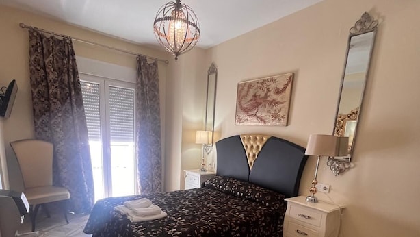 A bedroom with a bed, a nightstand, a chair, a window with curtains, and a chandelier. - Hostal Flor de Lis - Lojo (Conil de la Frontera)