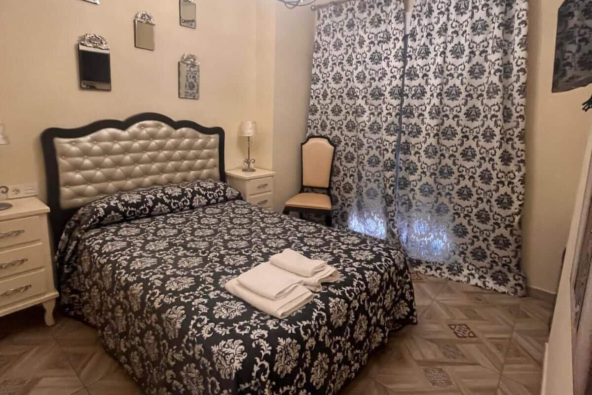 A bedroom with a tufted headboard bed, a bedside table with a lamp, and a patterned curtain.
