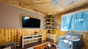 A cozy cabin interior with a wooden ceiling, a flat-screen TV, a sofa, a coffee table, and a window with a view of trees. - Pinecrest Family Summer Retreat Kid Playroom and Game Room (Island Park)