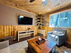 Een gezellig cabin interieur met houten plafond, een flatscreen televisie aan de muur, een comfortabele fauteuil, een bijzettafeltje en een raam met uitzicht op bomen.