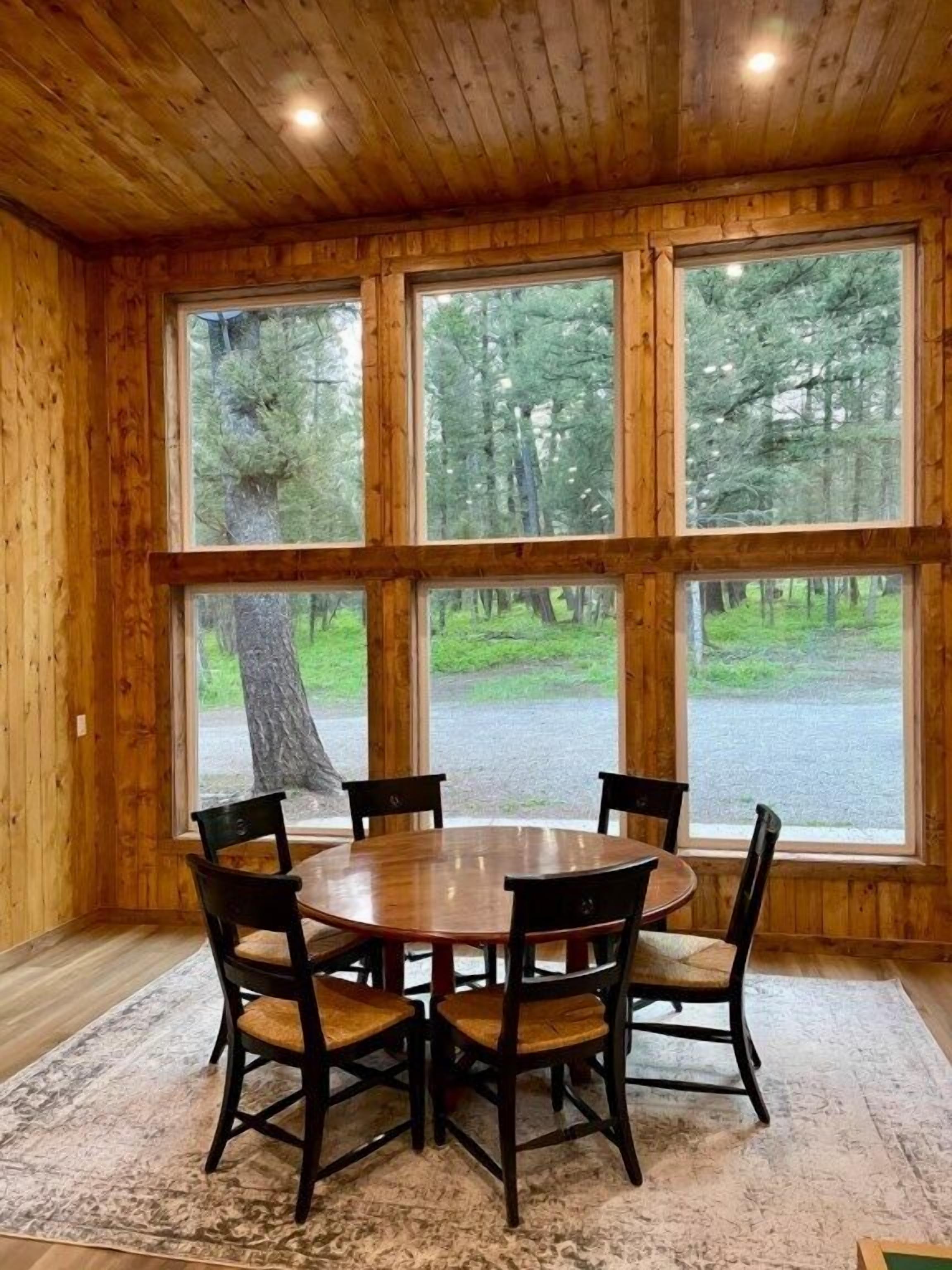 A wooden interior with a round table and chairs, large windows, and a view of a forest.