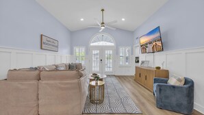 Ein geräumiges Wohnzimmer mit einem beigefarbenen Ecksofa, einem blauen Sessel, einer Holzkommode und einem großen Fernseher an der Wand.