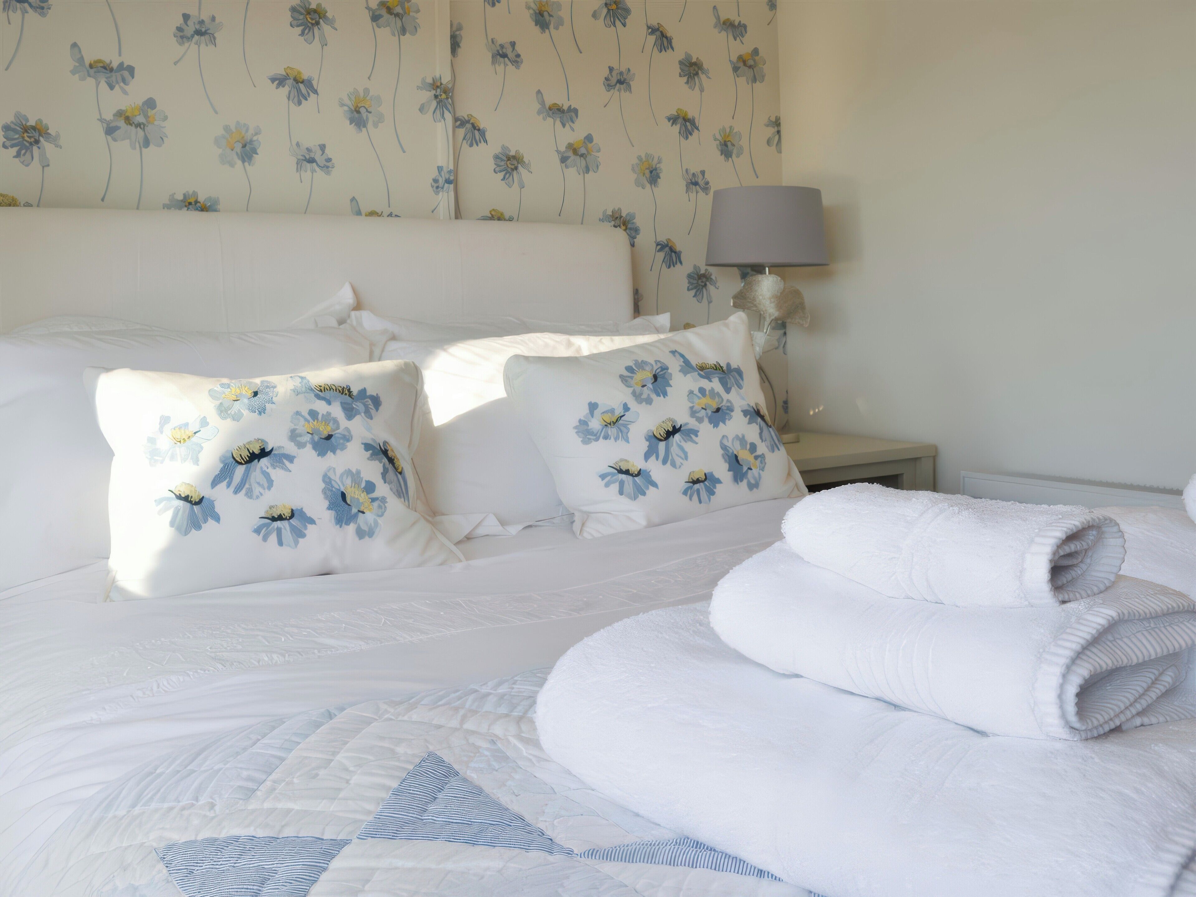 A neatly made bed with floral pillows, a bedside lamp, and folded towels.