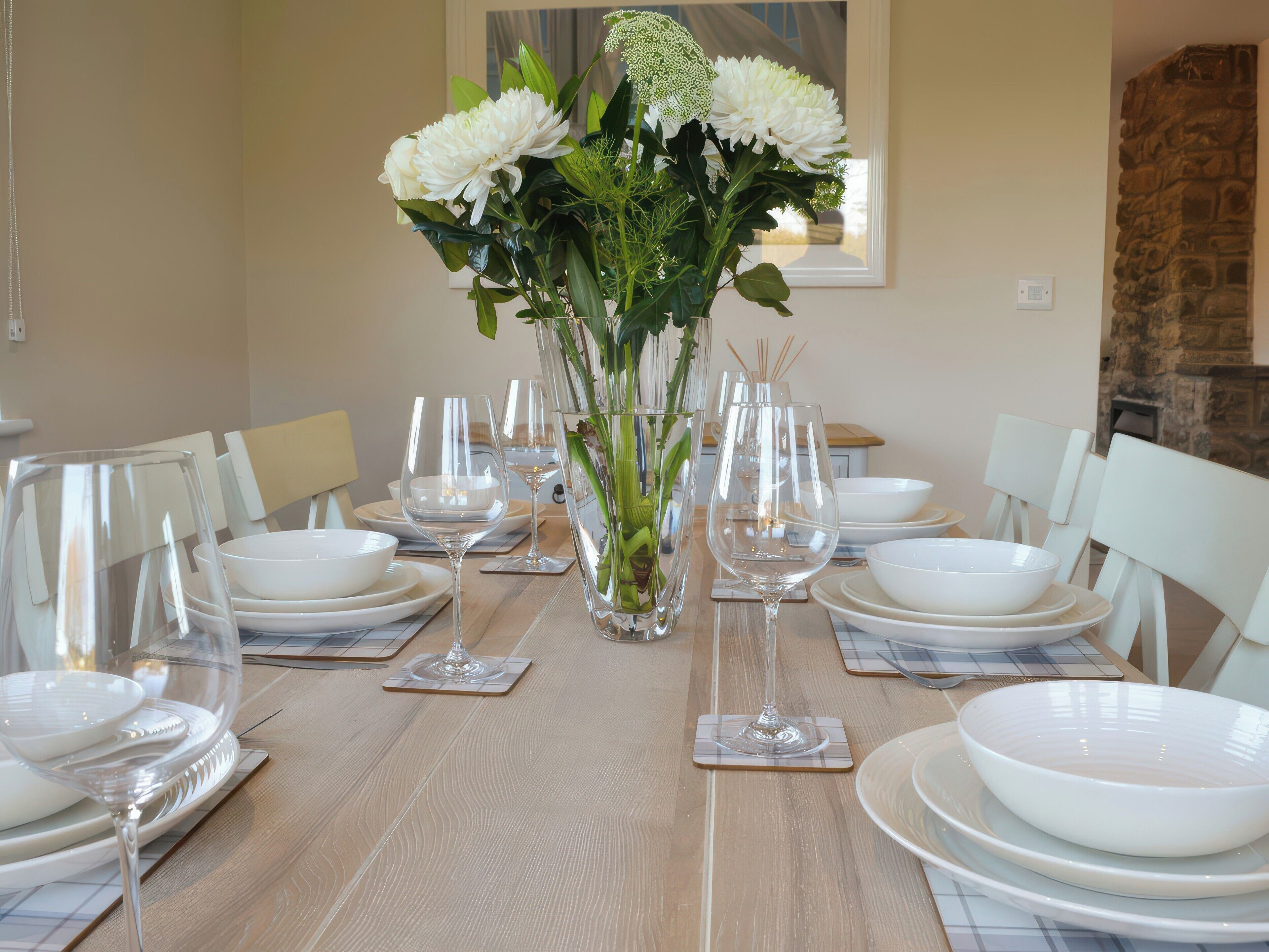 A dining table set with white plates, bowls, and wine glasses, centered with a vase of flowers.