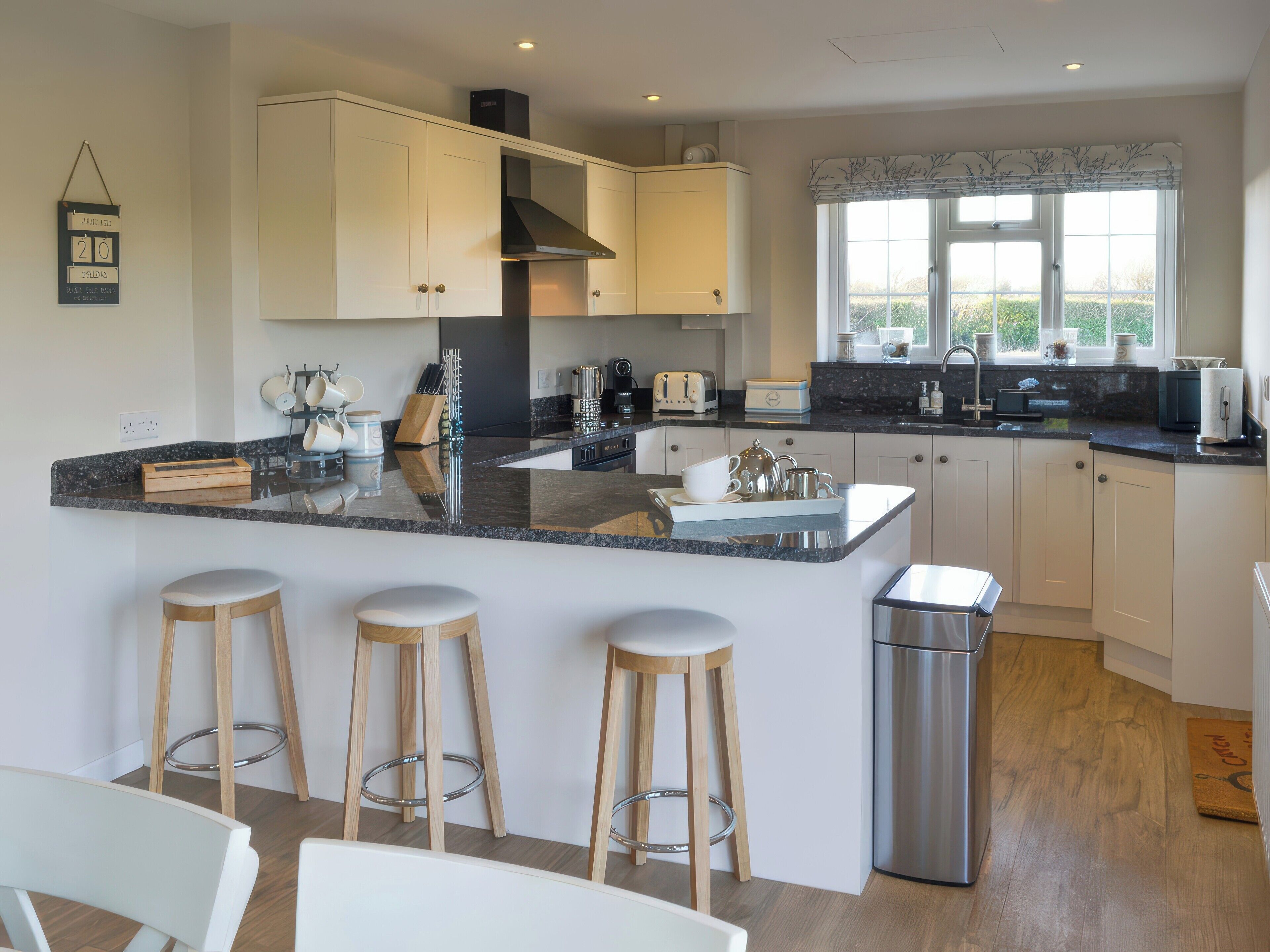 A modern kitchen with white cabinets, a granite countertop, and a stainless steel sink. There are three wooden stools and a trash bin.