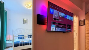 Una habitación moderna con un televisor de gran tamaño montado en la pared mostrando una interfaz de juego, una cama con estructura blanca y suelo de madera.