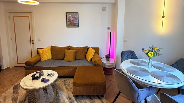 Una sala de estar moderna con sofá, reposapiés, mesa de centro y una mesa de comedor con sillas. Hay un cuadro en la pared y un jarrón con flores sobre la mesa de comedor.