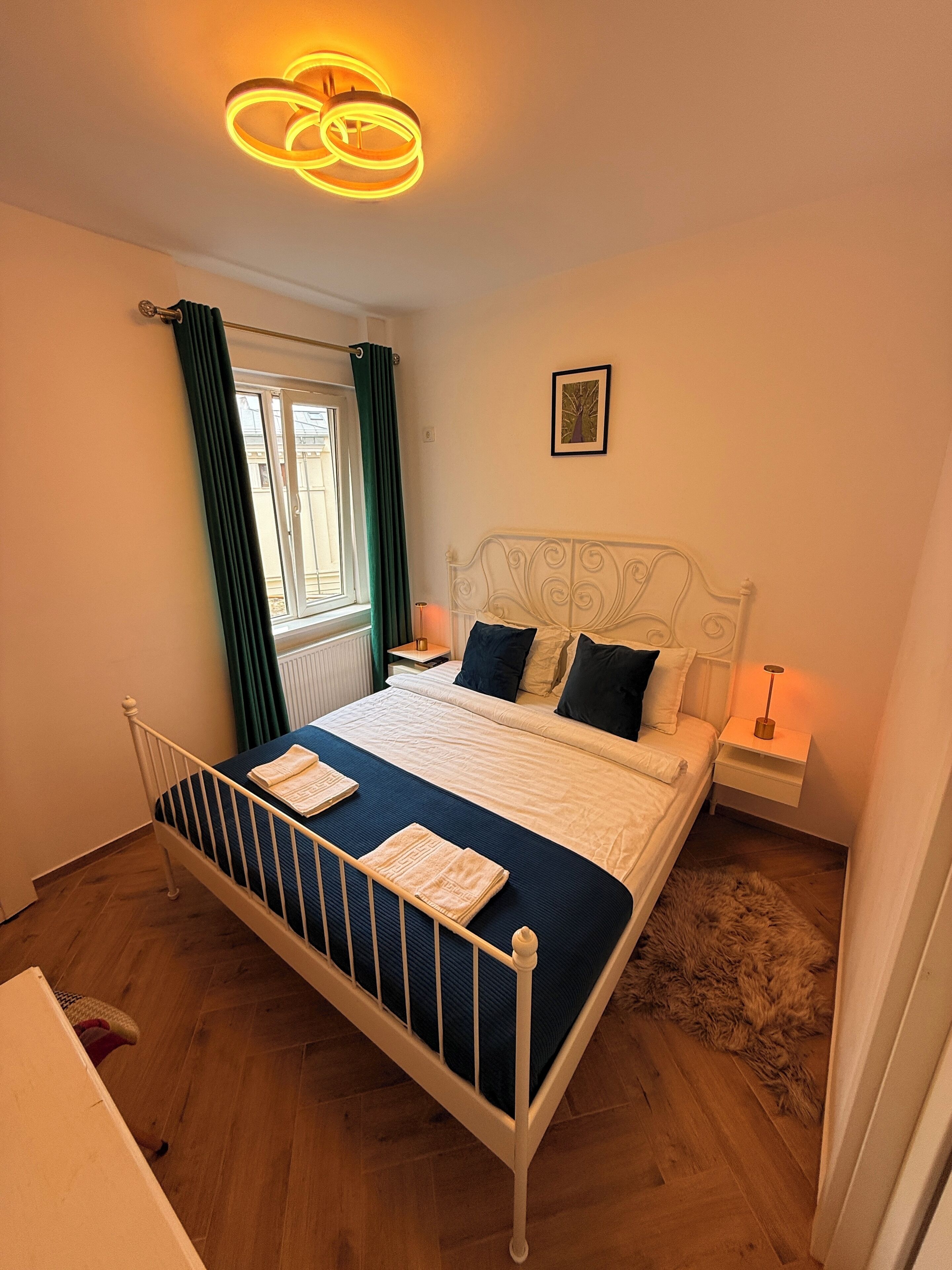 Un dormitorio ordenado con cama, mesitas de noche, una ventana con cortinas, un cuadro enmarcado en la pared y una lámpara en el techo.