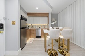 A modern kitchen with white cabinets, a stainless steel refrigerator, and a dishwasher. There is a dining area with white chairs and gold stools. - Orlando 2BR Apt, Full Kitchen, Near Epic Universe (Orlando)