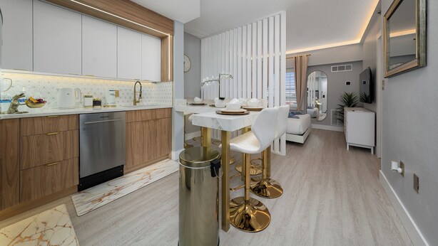 Een moderne keuken met witte kasten, een roestvrijstalen vaatwasser en een eethoek met een witte tafel en gouden stoelen.