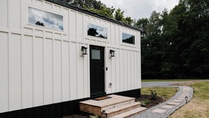 A white building with a black door, two windows, and a wooden staircase leading to the entrance. - Shadow’s Hideaway - Tiny Home w/ Fire Pit + Views (Cleveland)