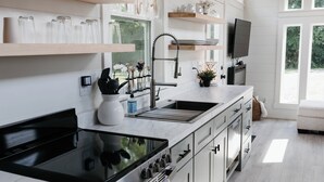 A modern kitchen with a wooden ceiling, large windows, and a TV mounted above the sink. - Shadow’s Hideaway - Tiny Home w/ Fire Pit + Views (Cleveland)
