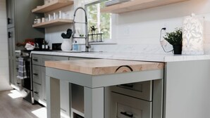 A kitchen with a central island, wooden shelves above, and a window providing natural light. - Shadow’s Hideaway - Tiny Home w/ Fire Pit + Views (Cleveland)