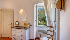 Una habitación con un tocador blanco, un espejo, una cesta de mimbre y una silla junto a una ventana con vistas a una colina.