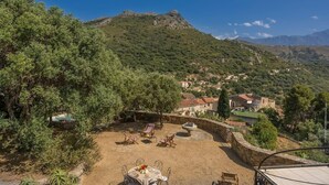 Un patio con mobiliario exterior y una mesa lista para comer, rodeado de vegetación y con vistas a un pueblo en la ladera.