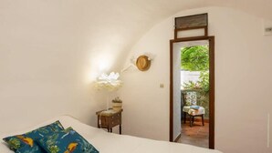 Un dormitorio con cama, una mesita de noche con lámpara, un sombrero colgado en la pared y vistas a un patio exterior.
