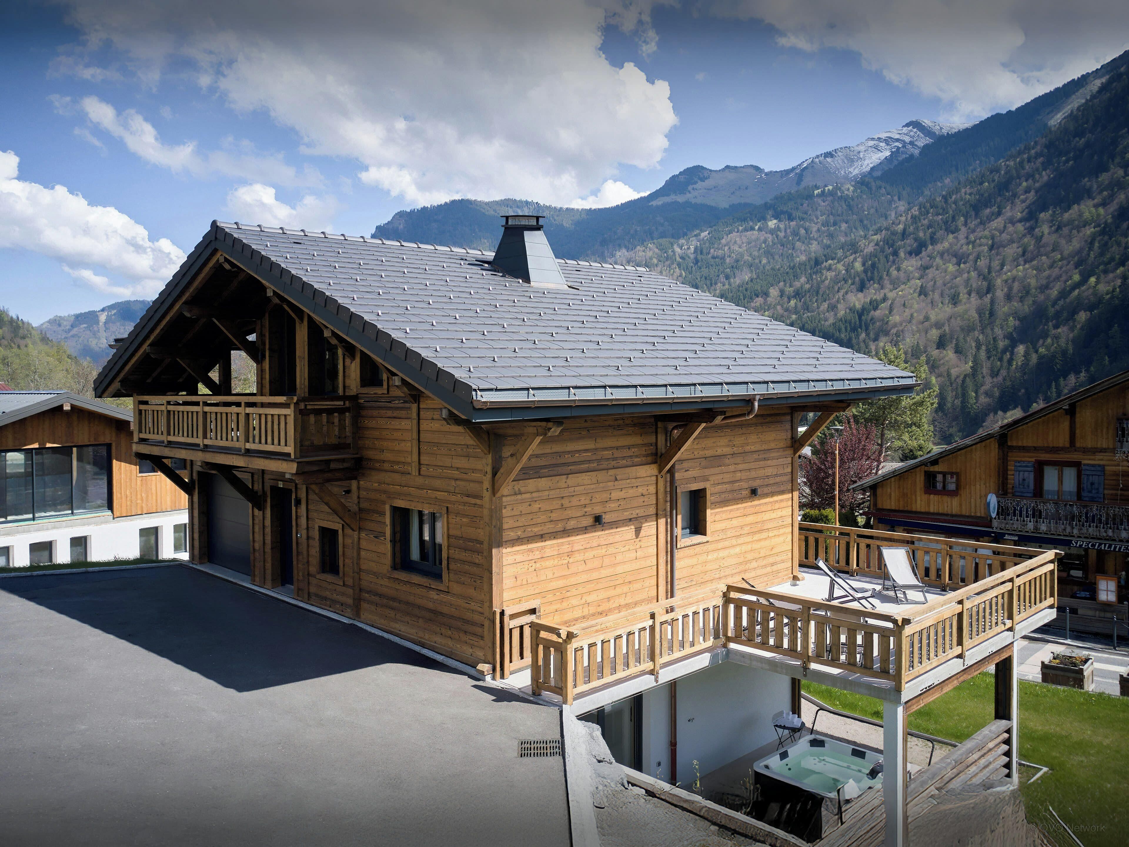 A wooden chalet with a balcony, a hot tub, and a mountain backdrop.