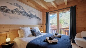 Quarto de cabana de madeira com cama, almofadas, abajur e uma janela grande com vista para montanhas.