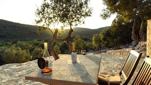 A stone table with a bottle, glasses, and a candle, surrounded by wooden chairs, set against a scenic hillside backdrop. - Luxurious home in Sali with WiFi (Sali)
