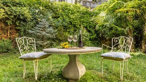 Un jardín con una mesa redonda y dos sillas, una botella de vino y un ramo de flores.