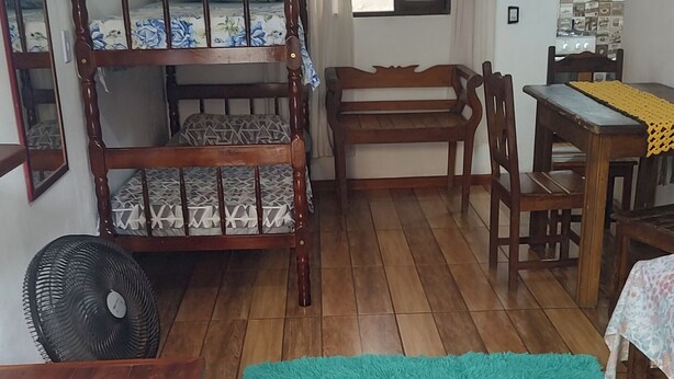 A room with a wooden bunk bed, a wooden desk, a chair, a small table, a fan, and a window with curtains. - Suíte Familiar em Paraty  (Paraty)