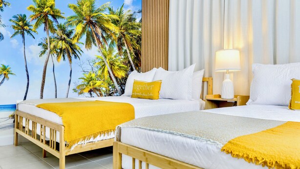 A hotel room with two beds, a view of palm trees, a wooden headboard, a white bedspread, and a yellow throw blanket.