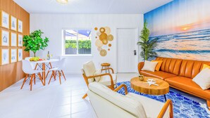 A modern living room with a white sofa, a dining table with chairs, a coffee table, and a large painting of a beach scene.