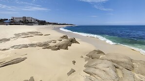 Uma praia com pedras grandes em primeiro plano, céu azul límpido e um edifício com vários andares ao fundo.