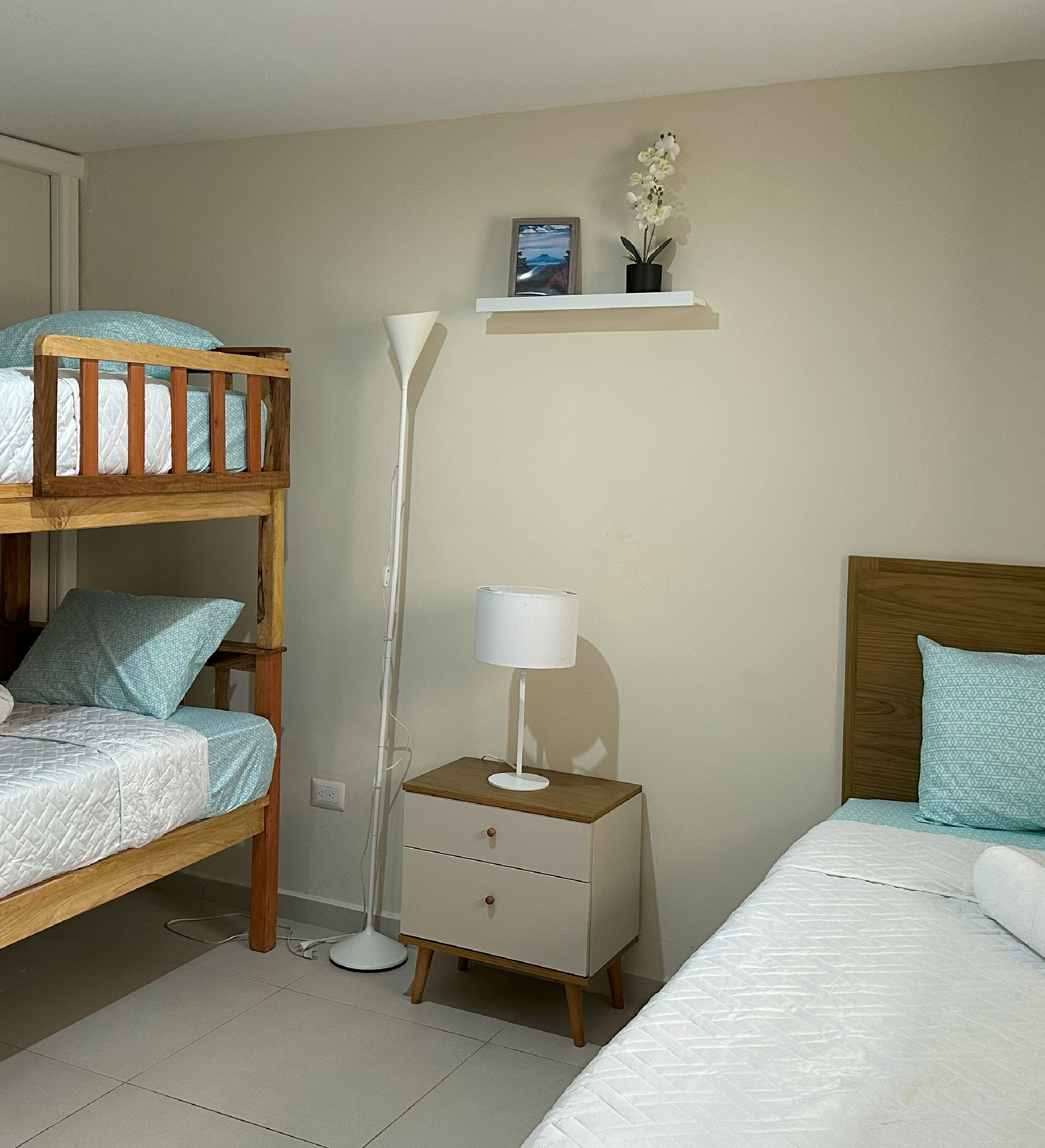 Um quarto com uma cama beliche, uma mesa de cabeceira com abajur, um criado-mudo e uma estante com uma foto e flores.