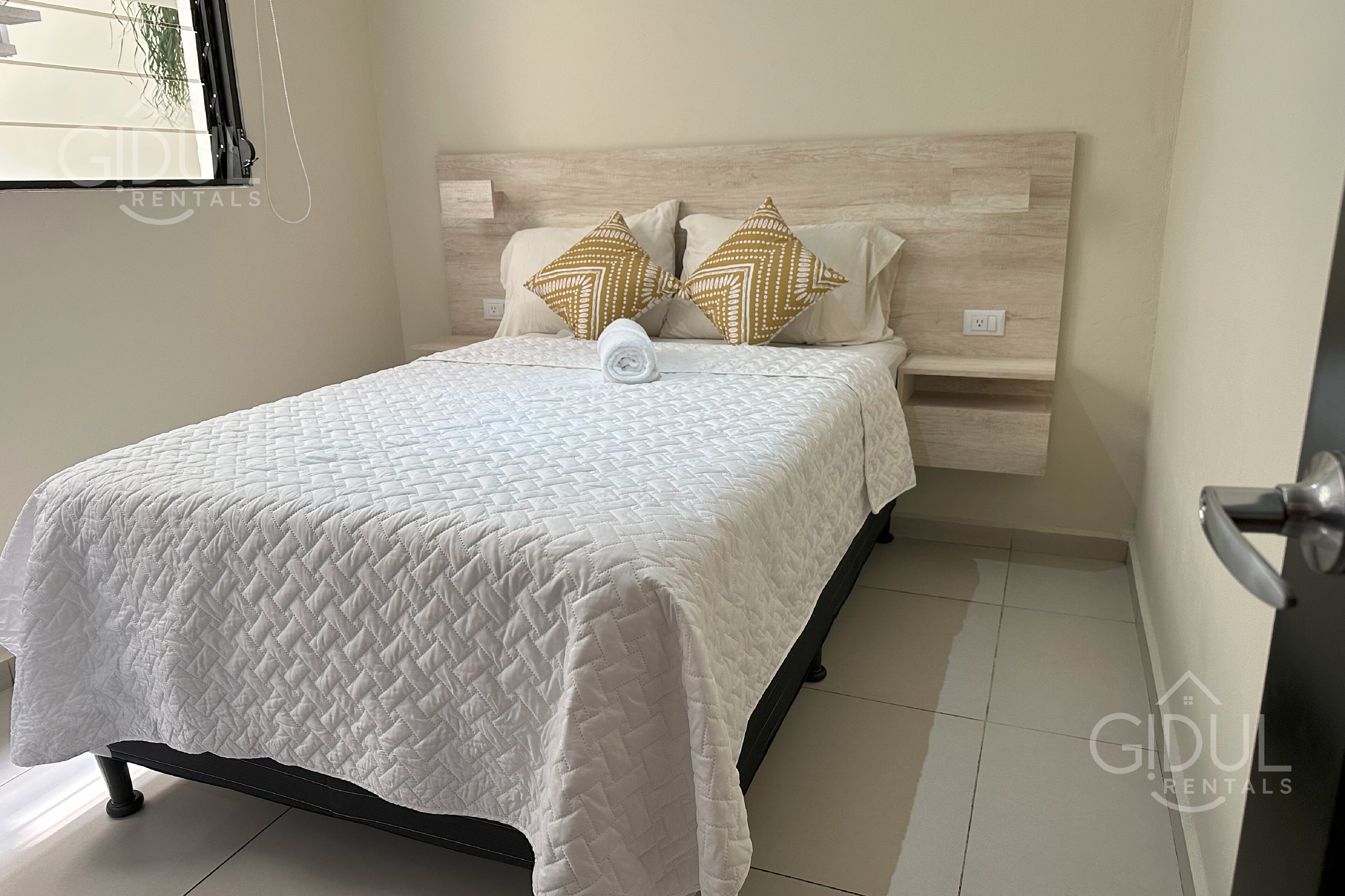 A neatly made bed with a white quilt, two decorative pillows, and a small bedside table with a light switch.