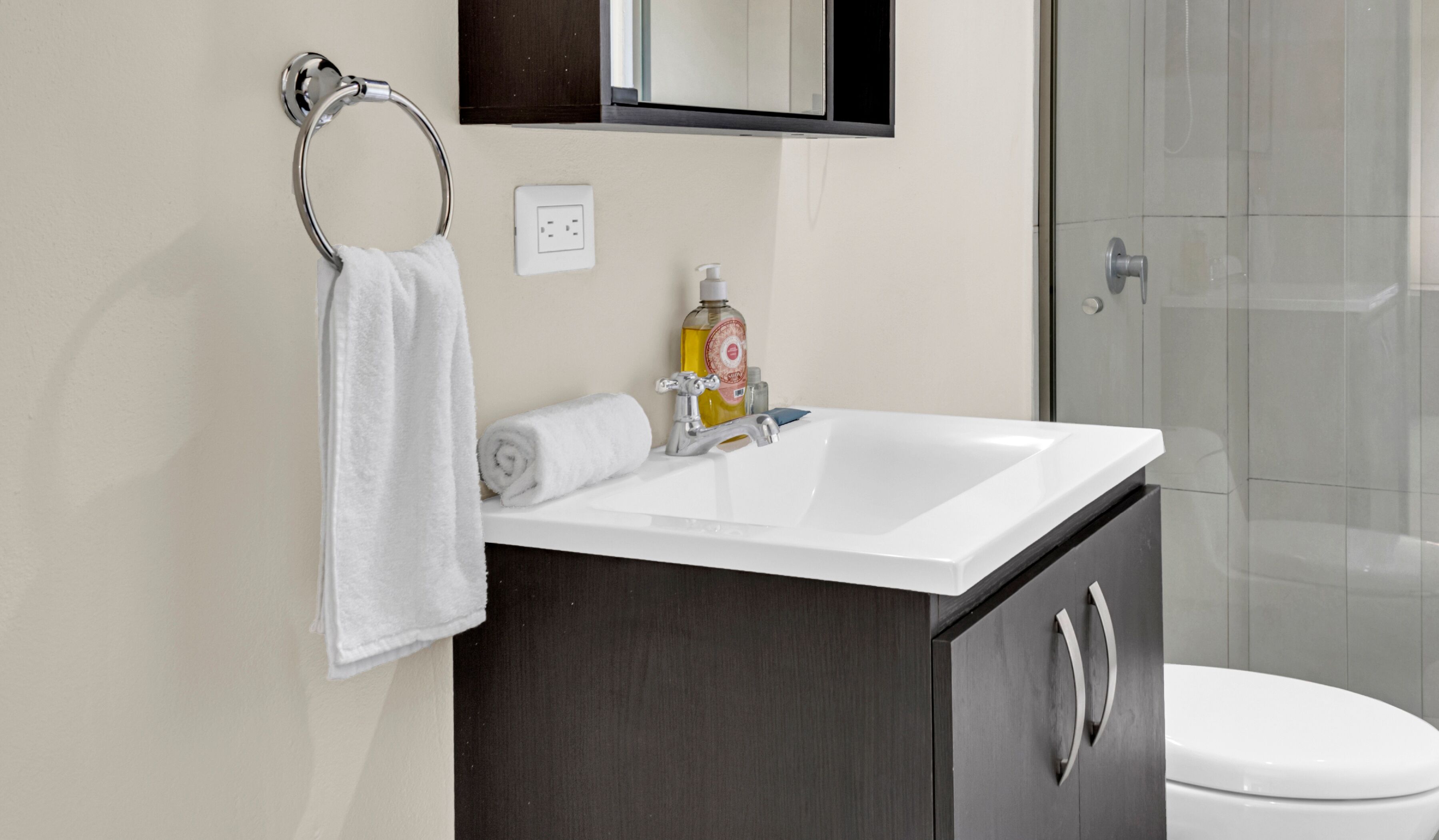 A bathroom with a white sink, dark wooden cabinet, a towel ring, a mirror, and a toilet.
