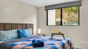 A bedroom with a bed, pillows, a quilt, a bedside table with a lamp, and a window with a view of greenery.