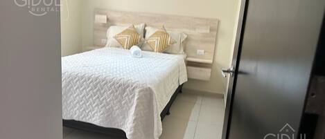 A neatly made bed with a white quilt and two decorative pillows, a wooden headboard, and a small bedside table.
