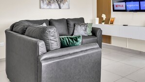 A grey leather sofa with multiple cushions, a white side table with a framed picture, and a wall-mounted TV displaying streaming services.