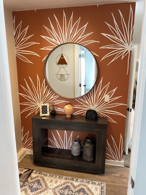 A hallway with a brown wall featuring white leaf patterns, a round mirror above a dark wooden console, and a patterned rug on the floor. - Cozy studio in Washington with WiFi, AC, close to restaurants & shopping. (Washington)