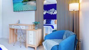 Une chambre avec un bureau en bois, un lit doté d’une tête de lit bleue, un téléviseur fixé au mur et une lampe de chevet.