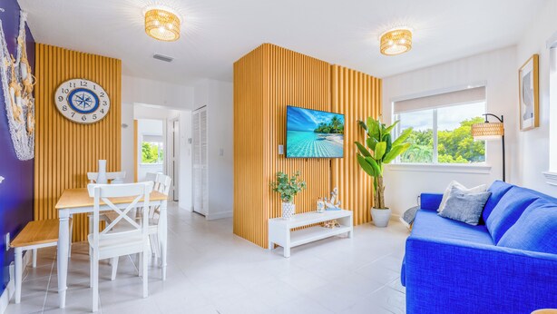 Un salon moderne avec un canapé bleu, une table à manger blanche et des chaises, un téléviseur fixé sur un mur en panneaux de bois, et une grande fenêtre donnant sur un paysage verdoyant.