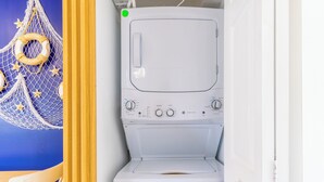 Une petite machine à laver blanche, placée dans une petite buanderie aux murs bleus, avec une chaise en bois.