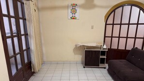 Ein Zimmer mit Fliesenboden, einem dunkelbraunen Sofa, einer kleinen Holzkabine und einem Fenster mit Zierrahmen.
